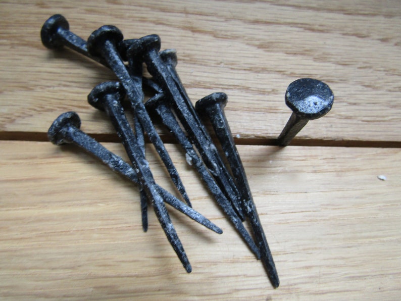 May include: A group of ten black metal nails with pointed tips, lying on a wooden surface. One nail is standing upright.
