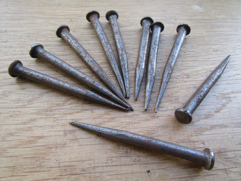 May include: A collection of ten rusty metal nails with pointed tips. The nails are arranged in a radial pattern on a wooden surface.