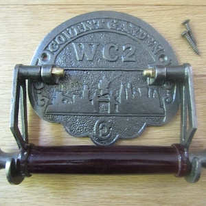 May include: A cast iron toilet paper holder with a decorative plaque featuring the text "Covent Garden WC2" and a silhouette of the London skyline. The holder has a dark brown wooden roller and two brass accents.