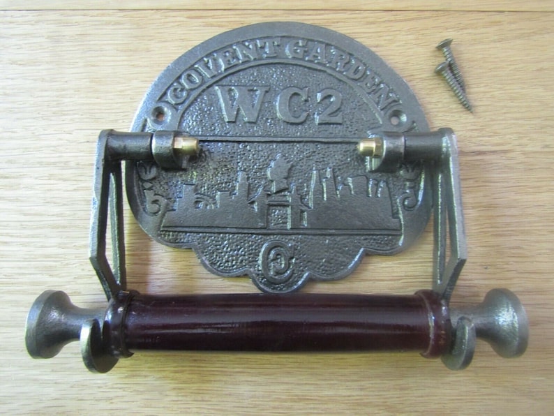 May include: A cast iron toilet paper holder with a decorative plaque featuring the text "Covent Garden WC2" and a silhouette of the London skyline. The holder has a dark brown wooden roller and two brass accents.