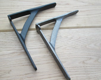 Puede incluir: Dos soportes de estante de metal, uno negro y otro plateado, se muestran sobre un fondo de madera clara. Los soportes están inclinados y tienen un diseño simple e industrial.