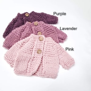 May include: Three knitted baby cardigans in shades of purple, lavender, and pink. Each cardigan has a button closure and is made with a soft, textured yarn.