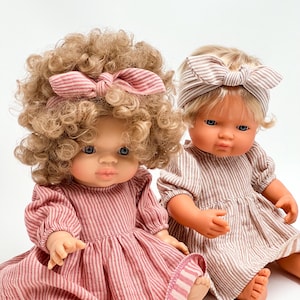 Op de afbeelding: Twee poppen die gestreepte jurken en hoofdbanden dragen. De pop links heeft krullend blond haar en draagt een roze-wit gestreepte jurk en hoofdband. De pop rechts heeft recht blond haar en draagt een bruin-wit gestreepte jurk en hoofdband.