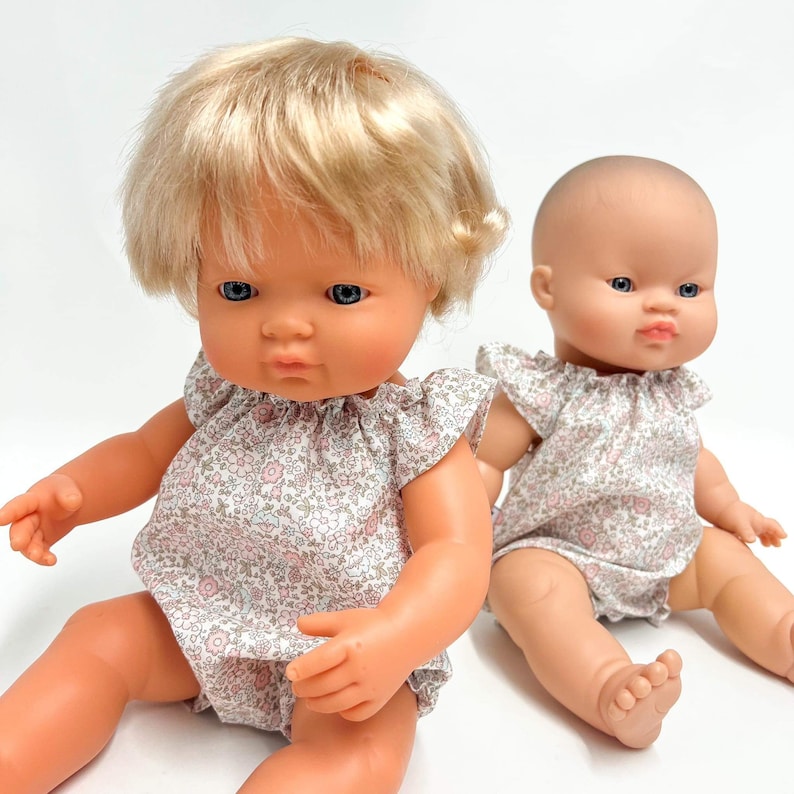 May include: Two dolls wearing matching floral print dresses. The doll on the left has blonde hair and blue eyes. The doll on the right has a bald head and blue eyes.