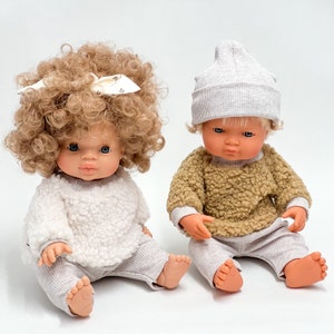 May include: Two dolls wearing fluffy sweaters and ribbed trousers. One doll has curly brown hair and a white bow. The other doll has a grey beanie and blonde hair.