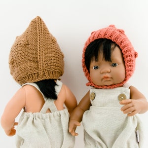 May include: Two dolls wearing knitted hats and dungarees. One doll has a brown knitted hat and the other doll has a pink knitted hat. Both dolls are wearing beige dungarees.