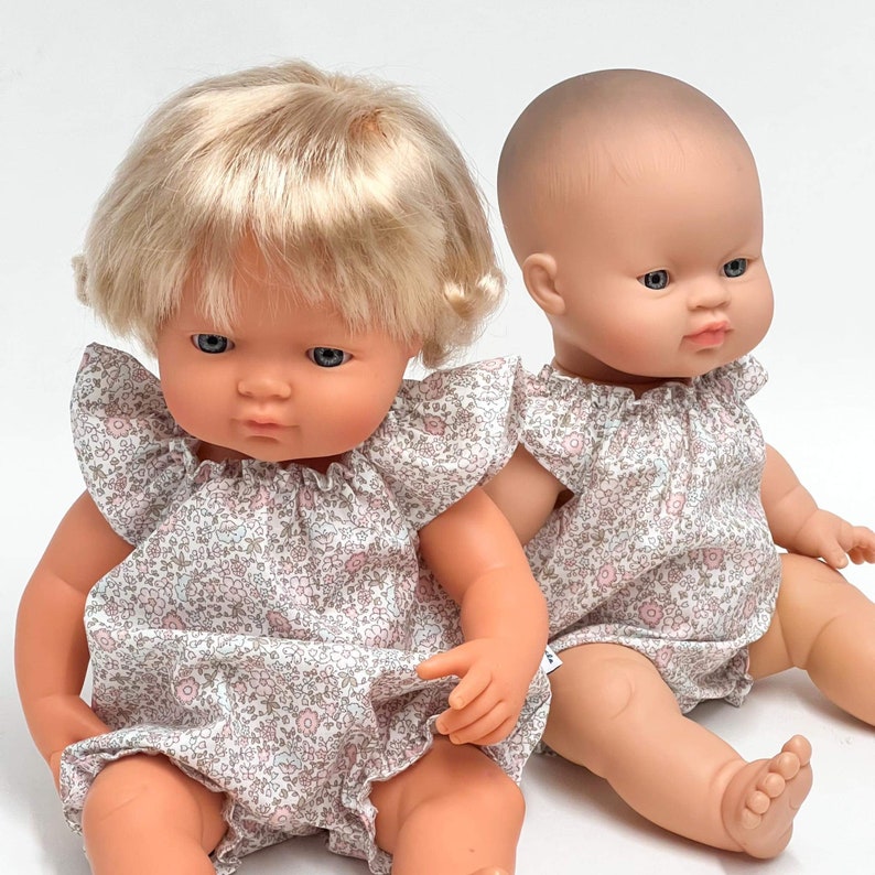 May include: Two baby dolls wearing matching floral print dresses. The doll on the left has blonde hair and blue eyes. The doll on the right has brown hair and brown eyes.