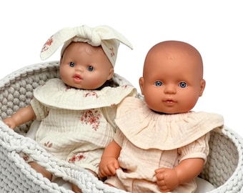 May include: Two baby dolls with blue eyes in a gray crocheted basket. One doll wears a cream-colored romper with a floral print and a matching headband. The other doll wears a solid cream-colored romper with a ruffled collar.