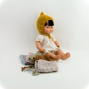 Puede incluir: Una muñeca con un gorro de ganchillo amarillo se sienta sobre una pila de gorros de ganchillo de varios colores, incluyendo gris, marrón, verde y rosa.