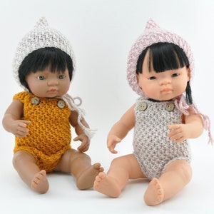 May include: Two knitted doll outfits with matching hats. One outfit is a mustard yellow romper with a white hat. The other outfit is a light pink romper with a pink hat.