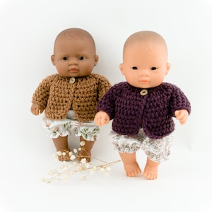 May include: Two dolls wearing crocheted cardigans. One doll has brown skin and is wearing a brown cardigan. The other doll has fair skin and is wearing a purple cardigan. Both dolls are wearing floral print bloomers.