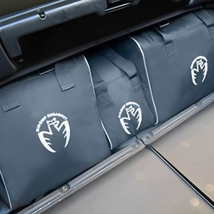 May include: Three gray fabric storage bags with white embroidered logos. The bags are in the trunk of a car.