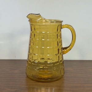 May include: Amber-colored glass pitcher with a textured, grid-like pattern. The pitcher has a handle and a spout for pouring. The design is reminiscent of vintage glassware. The pitcher is sitting on a wooden surface.