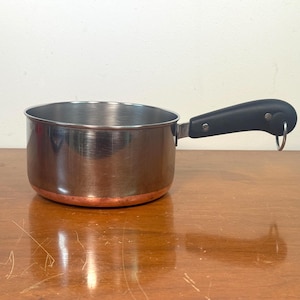 May include: A small, stainless steel saucepan with a copper-colored base and a black handle. The handle has a metal loop for hanging. The pan is sitting on a wooden surface.