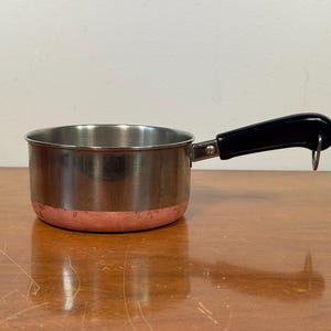 May include: A small stainless steel saucepan with a copper base and a black handle. The handle has a loop for hanging. The pan is on a wooden surface. The pan's diameter is approximately 15 cm.