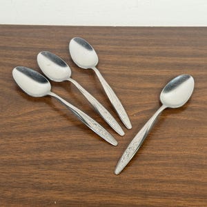 May include: Four stainless steel spoons with oval bowls and ornate floral designs on the handles. The spoons are arranged on a dark brown wooden surface, with the light reflecting off the polished metal.