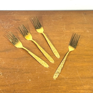 May include: A set of four gold-toned metal forks, each featuring a four-tine head and a handle decorated with an intricate, embossed floral pattern. The forks are arranged in a scattered formation, showcasing their metallic finish and design.