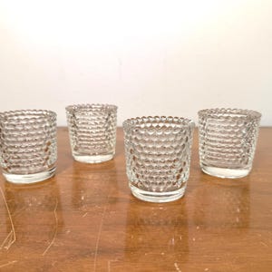 May include: A set of four clear glass votive candle holders, each featuring a distinct raised hobnail or bubble texture pattern on the exterior surface. The small, cylindrical vessels have a smooth rim and a thick, solid base for stability.