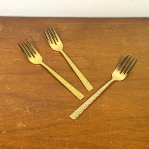 May include: Three gold-toned metal forks with four tines each. The handles feature a textured, stippled surface pattern that contrasts with the smooth, polished finish of the tines and the upper portion of the handle stems.