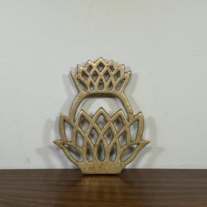 May include: A decorative, openwork metal thistle-shaped object in a warm gold tone. The design features a stylized flower with intricate cut-out details. The object is displayed on a dark wooden surface against a neutral background.