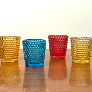 May include: Four coloured glass votive candle holders. The holders are in shades of yellow, blue, and red. Each has a textured, bubble-like pattern. The candle holders are on a brown surface.