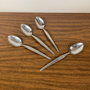 May include: Four silver spoons with rounded bowls and long, slender handles. The spoons are arranged in a fan shape on a brown wooden surface.