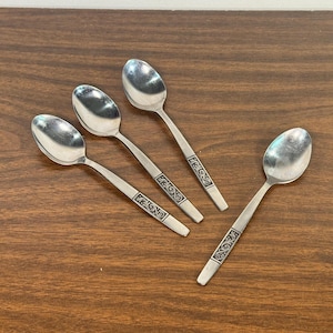 May include: Four silver spoons with ornate handle detailing are arranged on a dark wood surface. The spoons have a classic design, with a rounded bowl and a decorative pattern on the handle. The spoons are likely for serving or eating.