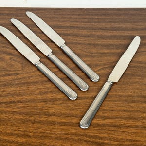 May include: Four silver butter knives with decorative handles are arranged on a dark wood surface. The knives have a classic, elegant appearance, with a shiny, reflective blade.
