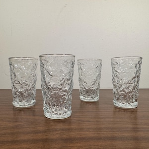May include: Four clear textured glass tumblers are arranged on a brown surface. The glasses have a unique, organic texture, resembling a bark-like pattern. The glasses are cylindrical and appear to be of varying heights, suitable for beverages.
