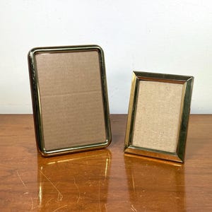 May include: Two gold-colored picture frames. One is rectangular and upright, the other is slightly angled. Both frames have a gold border and a cardboard backing. They are displayed on a wooden surface.