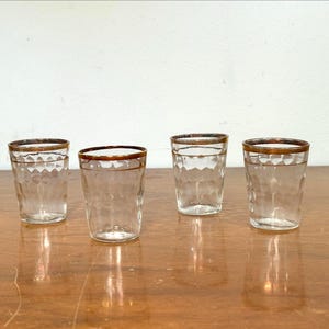 May include: Four clear drinking glasses with a textured, honeycomb-like pattern. Each glass has a gold-colored rim. The glasses are arranged on a wooden surface, reflecting the light. The glassware is suitable for serving beverages.