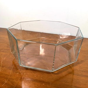 May include: Clear glass octagonal bowl. The bowl has a geometric design with eight sides. The glass is transparent, allowing a view of the interior. The bowl sits on a wooden surface.
