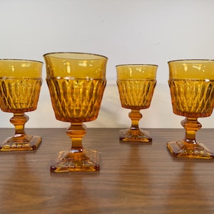 May include: Four amber-colored glass goblets with a diamond-shaped pattern. The goblets have square bases and are arranged on a wooden surface.