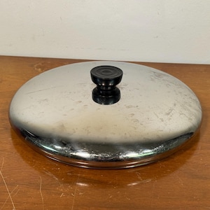 May include: A stainless steel pot lid with a black handle. The lid is round and has a shiny, reflective surface. The handle is a small, rounded knob. The lid is sitting on a wooden surface.