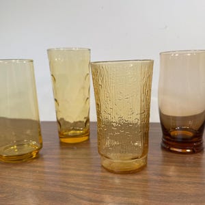 May include: Four amber glass drinking glasses with different textures. The glasses are on a wooden surface.