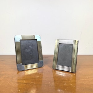 May include: Two vintage picture frames with a rectangular shape. The frames have a silver and gold color scheme with a dark interior. The frames are made of metal and are displayed on a wooden surface.