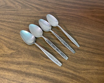 May include: Four stainless steel spoons with ornate floral and geometric designs on the handles. The spoons are arranged diagonally on a dark wood surface, showcasing their polished, reflective surfaces. The spoons are a classic silverware design.