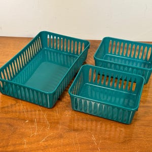 May include: Three teal plastic storage baskets of varying sizes are arranged on a wooden surface. The baskets have a rectangular shape with vertical slotted sides. The largest basket is rectangular, and the other two are square.
