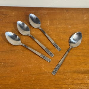 May include: Four stainless steel spoons with a decorative pattern on the handles are arranged on a wooden surface. The spoons have a classic design, with oval bowls and long, slender handles.
