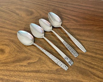 May include: Four silver-toned metal spoons arranged diagonally. Each spoon features a distinct embossed pattern on the handle, ranging from intricate floral scrollwork to geometric shapes and textured detailing on the stems.