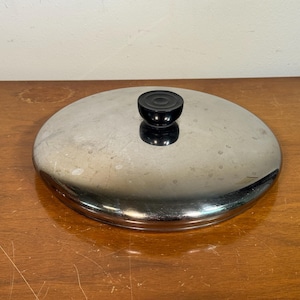 May include: A stainless steel pot lid with a black, round handle. The lid is circular and reflective, with a slightly domed shape. The handle is centered on the lid and appears to be made of a hard plastic material. The lid is sitting on a wooden surface.
