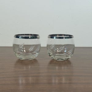 May include: Two clear, round drinking glasses with a silver rim and etched leaf design. The glasses are sitting on a brown wooden surface. The glasses are empty.