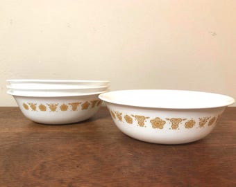May include: A set of four white ceramic bowls with a gold floral pattern around the rim. The bowls are stacked and arranged on a wooden surface, showcasing a vintage kitchenware design.