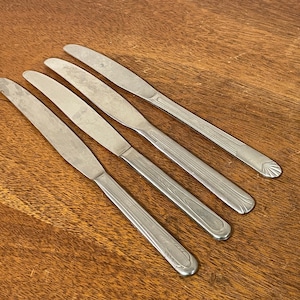 May include: Four silver dinner knives with ornate handles. The knives are laid out on a wooden surface.