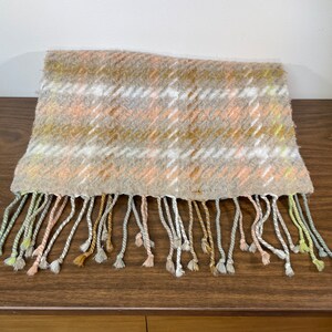 May include: A soft, beige, pink, and white plaid scarf with a fringe. The scarf is made of a fuzzy material and has a subtle sheen.