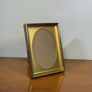 Peut inclure: Un cadre photo doré avec une ouverture ovale. Le cadre est légèrement incliné, posé sur une surface en bois. Le cadre a une bordure dorée et un dos marron.