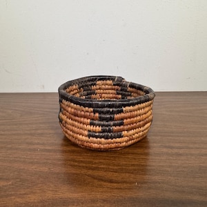 May include: A small, round woven basket with a geometric pattern. The basket is made of natural fibers in shades of brown and black. The basket is sitting on a dark wooden surface.