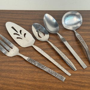 Peut inclure: Ensemble de couverts anciens en argent, comprenant une fourchette de service, une pelle à tarte, deux cuillères de service et une louche. Les couverts ont une finition argentée avec des motifs floraux sur les manches. Les pièces sont disposées sur une surface en bois.