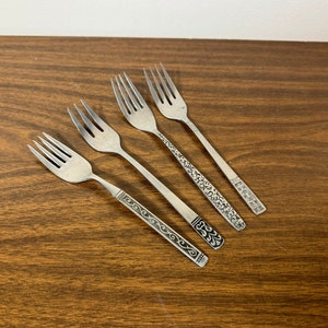 May include: Four silver forks with ornate designs on the handles. The forks are arranged in a row on a wooden surface.