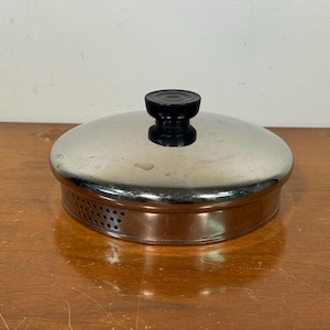 May include: A stainless steel pot lid with a black handle. The lid has a shiny, reflective surface and a copper-colored band around the edge with small ventilation holes. The lid is sitting on a wooden surface.